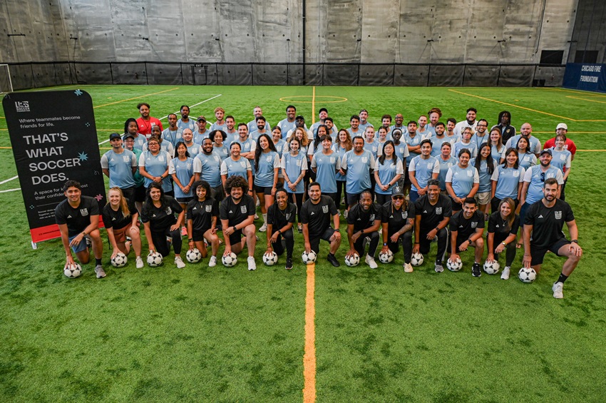 A group photo of about 70 individuals are lined up for a group photo on an indoor turf field. 15 of them are wearing black jerseys and are kneeling in one line in the front. the rest are the trainers who are standing in three rows and wearing light blue jerseys.